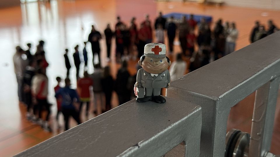 Zu sehen ist ein kleines DRK-Männchen, dass auf einem Geländer steht. Der Blick geht schräg nach unten in die Wehlehalle. Im Hintergrund ist unten unscharf eine Menschenmenge zu sehen.