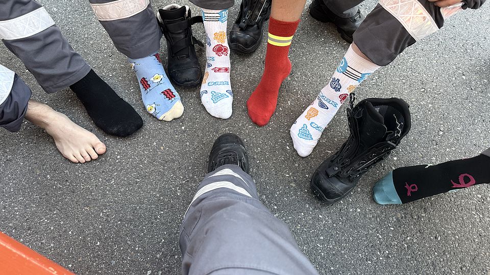 zu sehen sind 8 Füße. Von oben her im relativen Halbkreis sind: 1 Fuß ist nackt, 1 Fuß mit schwarzer Socke, 1 Fuß mit Motiv, das aussieht wie Speck und Spiegeleier, 1 Socke mir JRK-Motiven, 1 rote Socke mit 2 gelben Streifen und einem grauen Streifen zwischen den gelben oben am Bund, 1 weitere gleiche Socke mit JRK-Motiven, 1 ausgezogener Einsatzstiefel 1 Socke mit Schleifen-Motiven. Mittendrin von unten her ein Fuß mit Einsatzstiefel und grauer Einatzhose bis zum Knie.